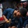 Hull Technicians Aboard USS Boxer Perform Welding, Grinding A young person wearing blue naval coveralls is using a hammer and chisel or punch on a metal piece secured in a bench vise. A second individual in similar blue U.S. Navy coveralls, with a partially visible name tag reading 'ELGARN', is assisting nearby. The setting appears to be a shipboard machine or repair shop with various tools and equipment visible in the background.