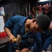 A young person wearing blue naval coveralls is using a hammer and chisel or punch on a metal piece secured in a bench vise. A second individual in similar blue U.S. Navy coveralls, with a partially visible name tag reading 'ELGARN', is assisting nearby. The setting appears to be a shipboard machine or repair shop with various tools and equipment visible in the background.