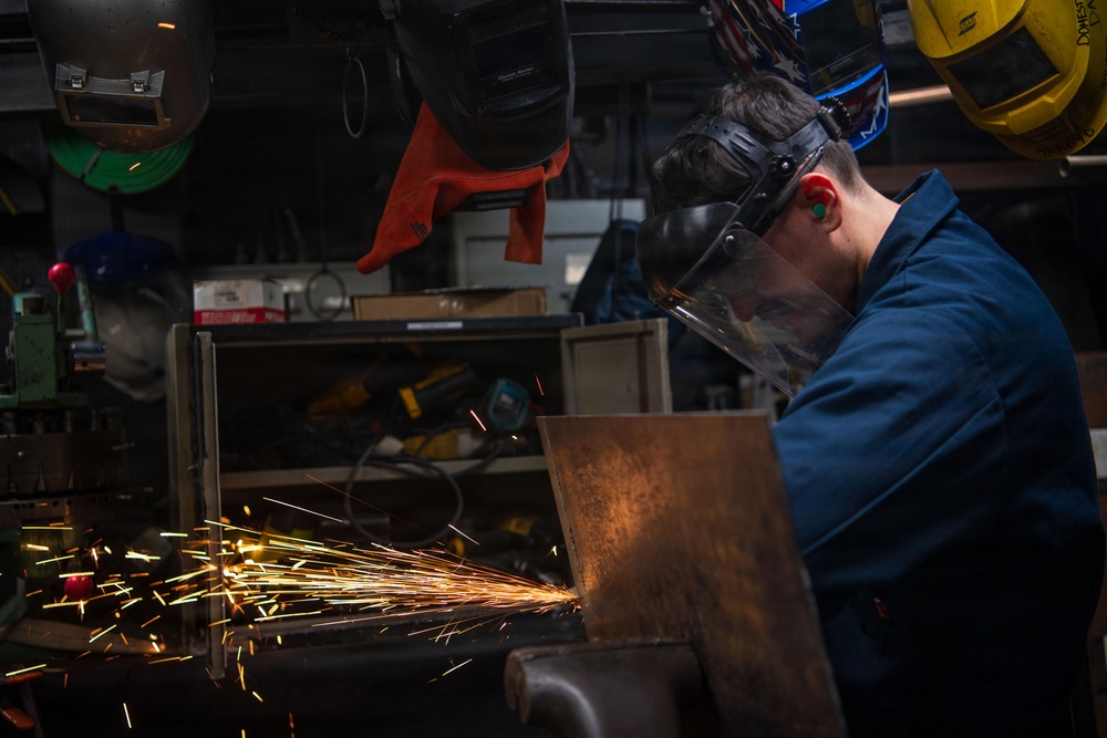A worker in blue coveralls is grinding or cutting metal in an industrial workshop, producing a dramatic shower of bright orange and yellow sparks. The worker is wearing a clear face shield pushed down over their face and has ear protection visible. The background shows a cluttered workshop environment with multiple welding helmets and various tools hanging or stored nearby.