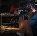 A worker in blue coveralls is grinding or cutting metal in an industrial workshop, producing a dramatic shower of bright orange and yellow sparks. The worker is wearing a clear face shield pushed down over their face and has ear protection visible. The background shows a cluttered workshop environment with multiple welding helmets and various tools hanging or stored nearby.