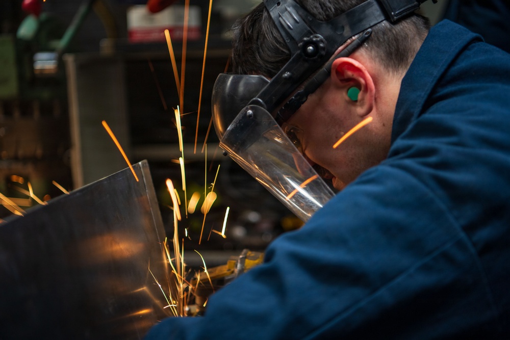 A worker is performing metal grinding or cutting on a flat metal sheet, producing a shower of bright orange and yellow sparks. The individual is wearing appropriate personal protective equipment including a face shield, hearing protection, and a blue work jacket. The image is captured in close-up, emphasizing the intensity of the sparks and the worker's focused posture.