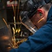 A worker is performing metal grinding or cutting on a flat metal sheet, producing a shower of bright orange and yellow sparks. The individual is wearing appropriate personal protective equipment including a face shield, hearing protection, and a blue work jacket. The image is captured in close-up, emphasizing the intensity of the sparks and the worker's focused posture.