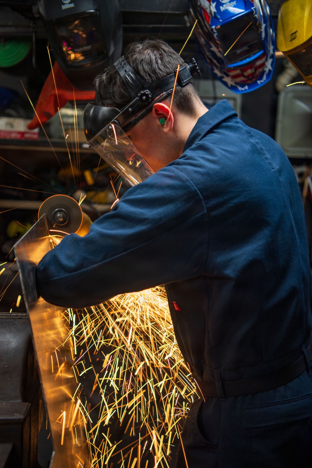 A worker in a blue coverall is using an angle grinder on a metal workpiece, generating a dramatic shower of bright orange and yellow sparks. The individual is wearing appropriate personal protective equipment including a clear face shield, hearing protection (green earplugs), and safety glasses. The background shows a workshop environment with multiple welding helmets hanging on the wall, including a Miller-branded patriotic American flag design helmet.