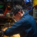 A worker in a blue coverall is using an angle grinder on a metal workpiece, generating a dramatic shower of bright orange and yellow sparks. The individual is wearing appropriate personal protective equipment including a clear face shield, hearing protection (green earplugs), and safety glasses. The background shows a workshop environment with multiple welding helmets hanging on the wall, including a Miller-branded patriotic American flag design helmet.