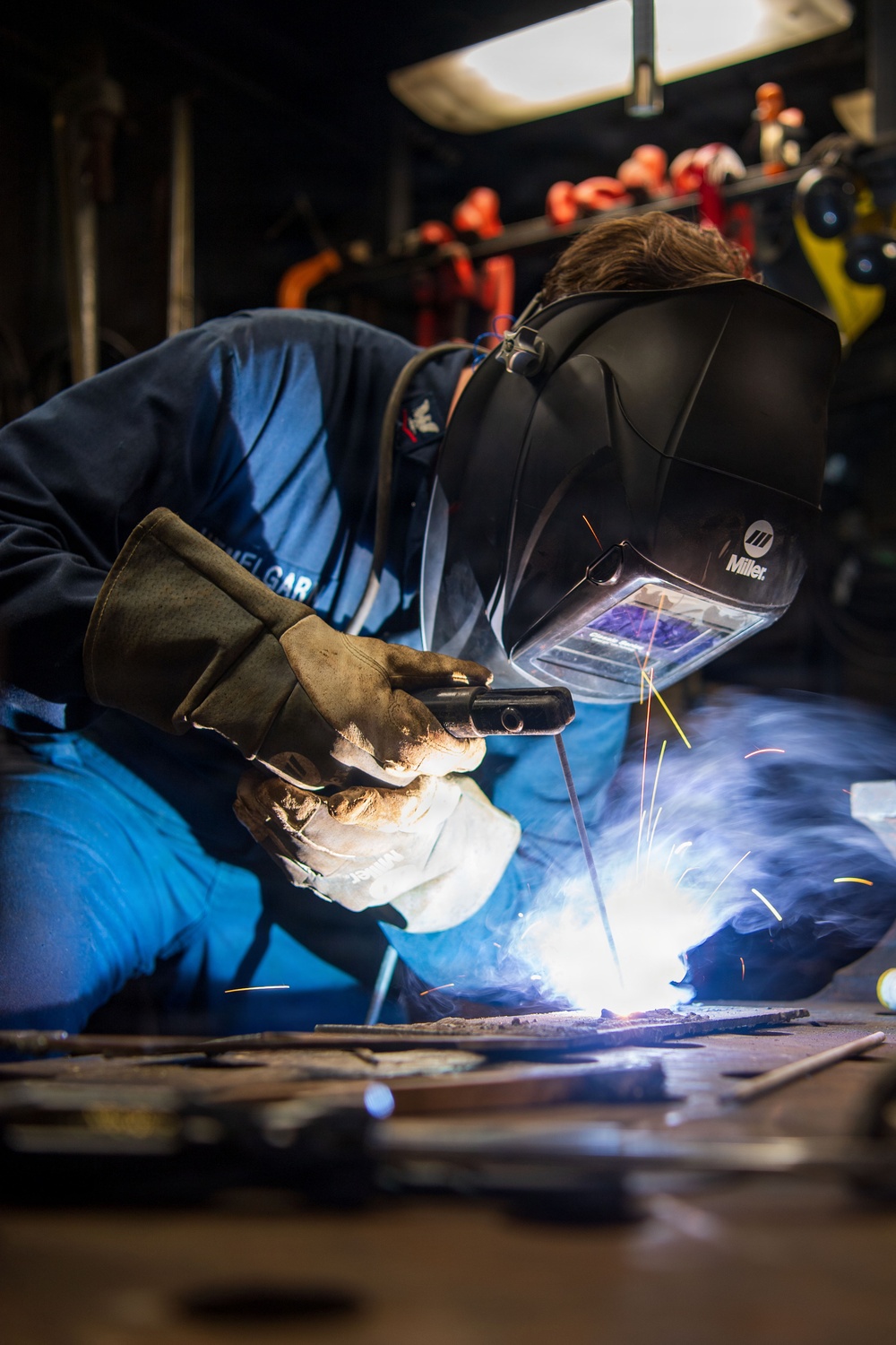A welder is actively welding metal pieces on a workshop floor, producing bright sparks, blue-white arc light, and smoke. The worker is wearing a Miller brand auto-darkening welding helmet and heavy leather protective gloves. The setting appears to be an industrial workshop with various tools and equipment visible in the background.