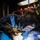 Hull Technicians Aboard USS Boxer Perform Welding, Grinding A welder is actively welding metal pieces on a workshop floor, producing bright sparks, blue-white arc light, and smoke. The worker is wearing a Miller brand auto-darkening welding helmet and heavy leather protective gloves. The setting appears to be an industrial workshop with various tools and equipment visible in the background.