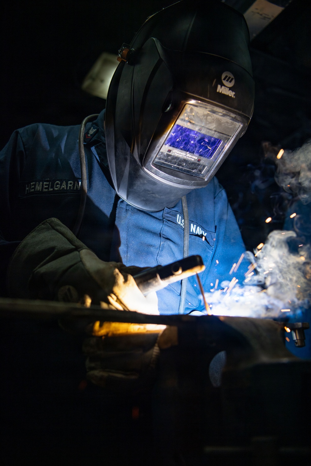 A U.S. Navy service member identified as 'HEMELGARN' is performing welding operations in a dark workspace. The welder is equipped with a Miller Classic Series auto-darkening welding helmet and protective gloves, actively welding metal components with visible sparks, smoke, and arc light. The dramatic lighting from the welding arc illuminates the scene against a dark background.