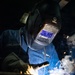 A U.S. Navy service member identified as 'HEMELGARN' is performing welding operations in a dark workspace. The welder is equipped with a Miller Classic Series auto-darkening welding helmet and protective gloves, actively welding metal components with visible sparks, smoke, and arc light. The dramatic lighting from the welding arc illuminates the scene against a dark background.