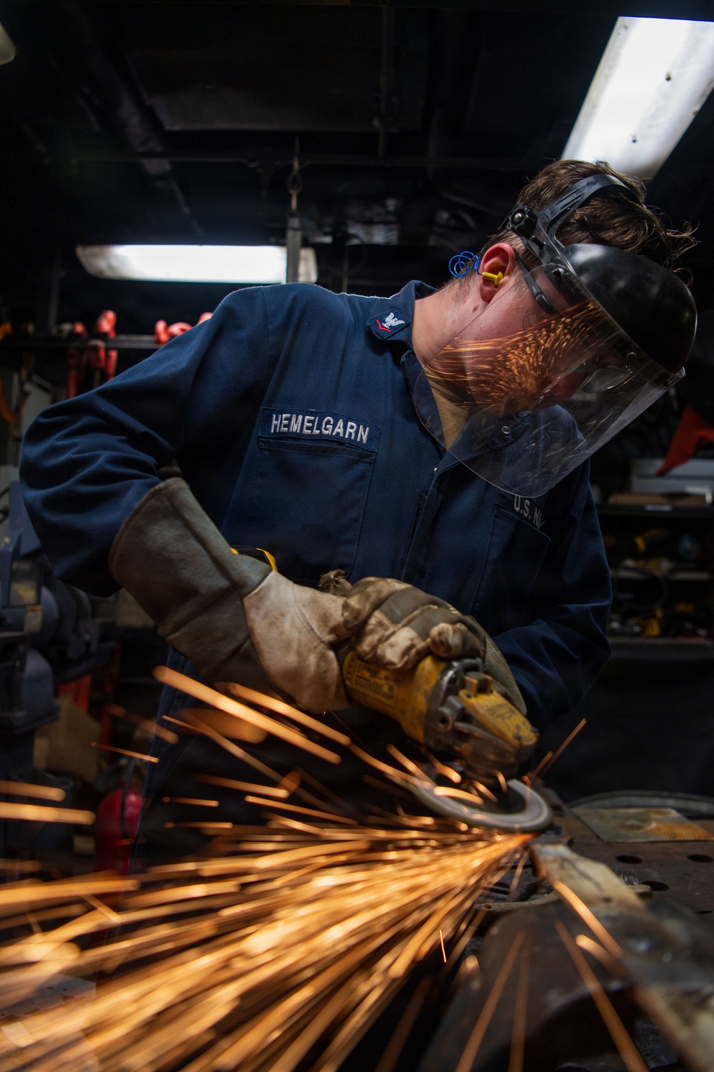 A U.S. Navy sailor named Hemelgarn is using an angle grinder on a metal surface in what appears to be a shipboard workshop or machine shop. Bright orange sparks are flying dramatically from the grinding operation. The sailor is wearing appropriate personal protective equipment including a face shield, safety glasses, ear protection, and heavy-duty work gloves.