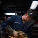 A U.S. Navy sailor named Hemelgarn is using an angle grinder on a metal surface in what appears to be a shipboard workshop or machine shop. Bright orange sparks are flying dramatically from the grinding operation. The sailor is wearing appropriate personal protective equipment including a face shield, safety glasses, ear protection, and heavy-duty work gloves.