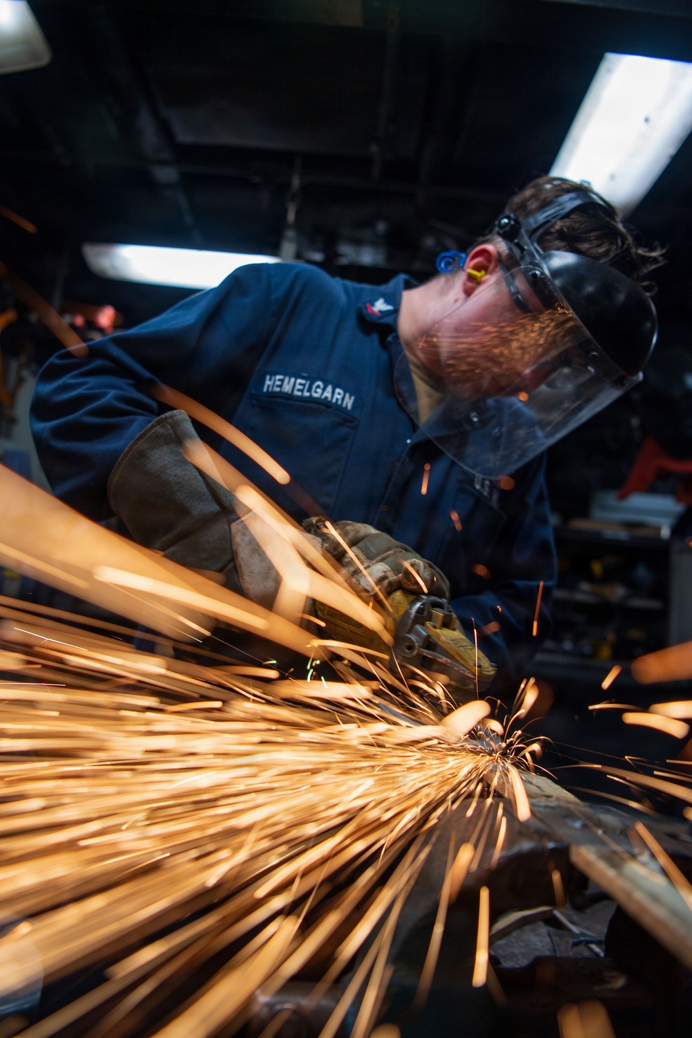 A person identified by name tape as 'HEMELGARN' is using an angle grinder on a metal surface, producing a dramatic shower of bright orange sparks. The individual is wearing a dark blue uniform consistent with U.S. military or naval service, along with appropriate personal protective equipment. The low-angle close-up shot emphasizes the intensity of the sparks flying toward the camera.