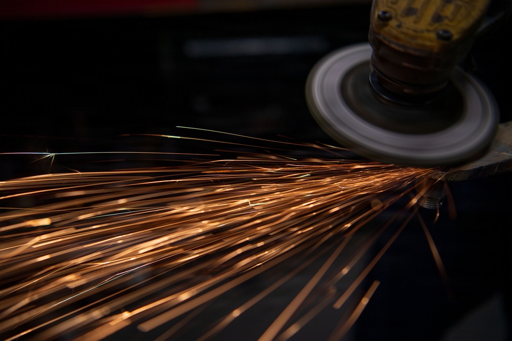 A close-up photograph of an angle grinder in operation, making contact with a metal surface and producing a dramatic shower of orange sparks. The sparks trail diagonally across the frame from right to left against a dark background, creating a visually striking industrial scene. The image captures motion blur on both the spinning grinding disc and the ejected spark trails.