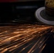A close-up photograph of an angle grinder in operation, making contact with a metal surface and producing a dramatic shower of orange sparks. The sparks trail diagonally across the frame from right to left against a dark background, creating a visually striking industrial scene. The image captures motion blur on both the spinning grinding disc and the ejected spark trails.