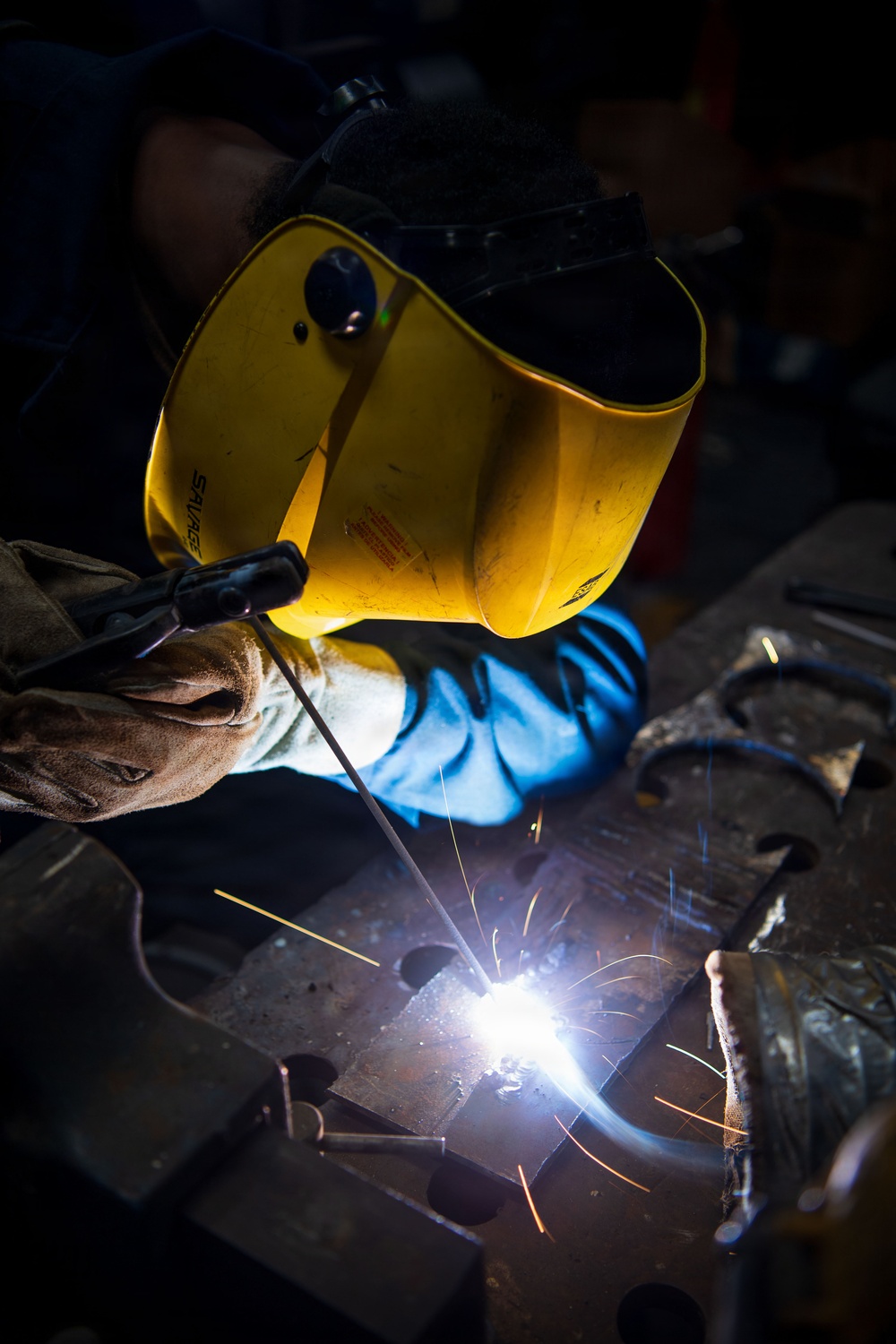 A welder is actively performing a welding operation on metal workpieces on a workbench. The welder is wearing a yellow 'Savage' branded auto-darkening welding helmet and protective gloves, with bright sparks and arc light visible at the weld point. The dramatic lighting from the welding arc illuminates the scene against a dark workshop background.
