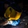 Hull Technicians Aboard USS Boxer Perform Welding, Grinding A welder is actively performing a welding operation on metal workpieces on a workbench. The welder is wearing a yellow 'Savage' branded auto-darkening welding helmet and protective gloves, with bright sparks and arc light visible at the weld point. The dramatic lighting from the welding arc illuminates the scene against a dark workshop background.