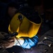 A welder is actively performing a welding operation on metal workpieces on a workbench. The welder is wearing a yellow 'Savage' branded auto-darkening welding helmet and protective gloves, with bright sparks and arc light visible at the weld point. The dramatic lighting from the welding arc illuminates the scene against a dark workshop background.