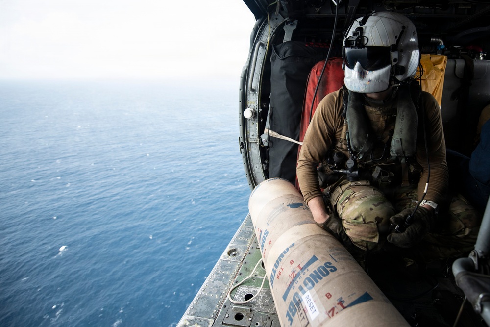 A military aircrew member in flight gear is seated at the open door of a helicopter over open ocean, preparing to deploy what appears to be a sonobuoy from a cylindrical tube. The operator is wearing a flight helmet with visor, multicam uniform, and survival vest while positioned at the edge of the aircraft. The image captures an active maritime patrol or anti-submarine warfare operation.