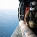 A military aircrew member in flight gear is seated at the open door of a helicopter over open ocean, preparing to deploy what appears to be a sonobuoy from a cylindrical tube. The operator is wearing a flight helmet with visor, multicam uniform, and survival vest while positioned at the edge of the aircraft. The image captures an active maritime patrol or anti-submarine warfare operation.