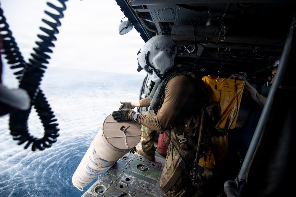A military helicopter crew member wearing a flight helmet and tactical gear is positioned at the open door of a helicopter over open ocean water. The crew member appears to be handling or deploying a cylindrical object, possibly a sonobuoy or supply canister, from the aircraft. At least one other crew member is visible inside the helicopter cabin holding what appears to be a yellow vest or equipment.