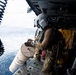 A military helicopter crew member wearing a flight helmet and tactical gear is positioned at the open door of a helicopter over open ocean water. The crew member appears to be handling or deploying a cylindrical object, possibly a sonobuoy or supply canister, from the aircraft. At least one other crew member is visible inside the helicopter cabin holding what appears to be a yellow vest or equipment.