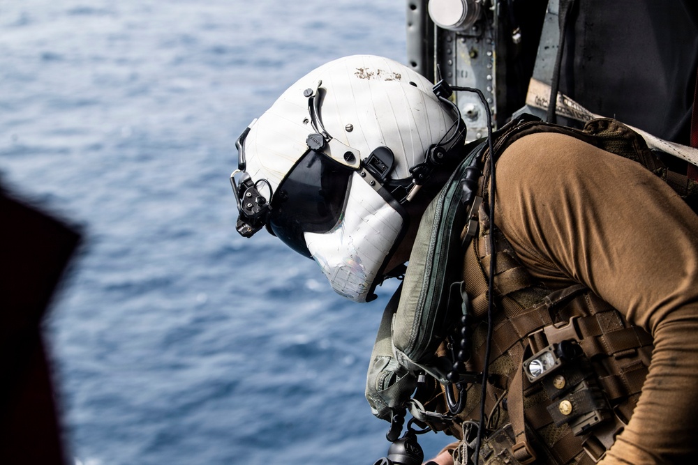 A military aircrew member or special operations personnel is positioned at the open door of a helicopter over open water. The individual is wearing a white flight helmet with a dark visor and is equipped with tactical gear including a plate carrier vest with attached equipment. The ocean is visible below, suggesting an airborne maritime operation.