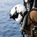 A military aircrew member or special operations personnel is positioned at the open door of a helicopter over open water. The individual is wearing a white flight helmet with a dark visor and is equipped with tactical gear including a plate carrier vest with attached equipment. The ocean is visible below, suggesting an airborne maritime operation.