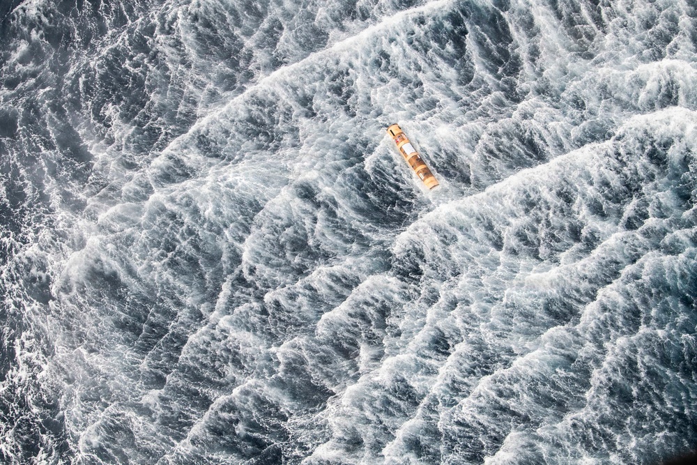 An aerial photograph captures a small vessel or life raft adrift in extremely rough, turbulent ocean waters. The sea surface is covered in large, chaotic white-capped waves suggesting severe storm conditions. The orange and yellow object appears tiny and vulnerable against the overwhelming scale of the churning water.