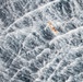 An aerial photograph captures a small vessel or life raft adrift in extremely rough, turbulent ocean waters. The sea surface is covered in large, chaotic white-capped waves suggesting severe storm conditions. The orange and yellow object appears tiny and vulnerable against the overwhelming scale of the churning water.