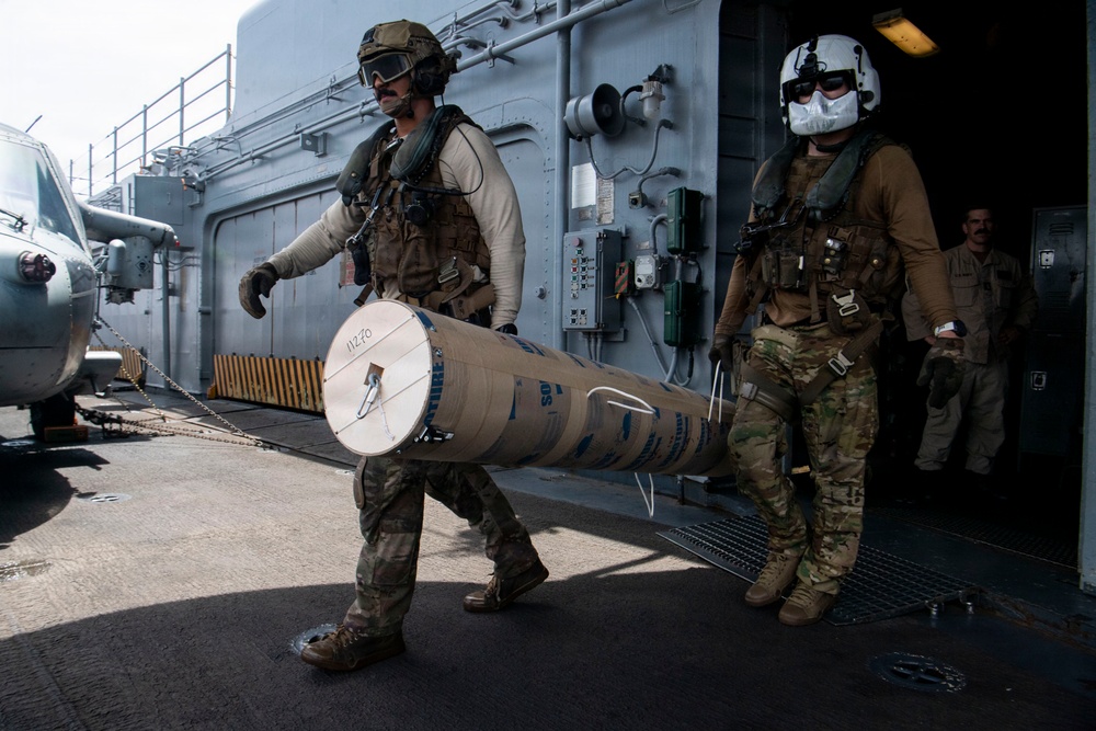 Two military personnel in full tactical gear are carrying a large cylindrical munition or ordnance container on the deck of a naval vessel. A third individual is partially visible in a doorway in the background. The setting appears to be the flight deck or exterior deck of a warship, with a helicopter partially visible on the left.