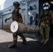 Two military personnel in full tactical gear are carrying a large cylindrical munition or ordnance container on the deck of a naval vessel. A third individual is partially visible in a doorway in the background. The setting appears to be the flight deck or exterior deck of a warship, with a helicopter partially visible on the left.