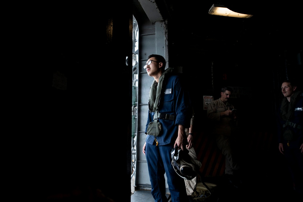 A military or naval personnel member stands near an open hatch or doorway in what appears to be a ship's interior, illuminated by natural light from outside. The individual is wearing a blue flight suit or coveralls with a survival vest and carrying a helmet. Two other personnel are visible in the darker background to the right.