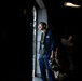 A military or naval personnel member stands near an open hatch or doorway in what appears to be a ship's interior, illuminated by natural light from outside. The individual is wearing a blue flight suit or coveralls with a survival vest and carrying a helmet. Two other personnel are visible in the darker background to the right.
