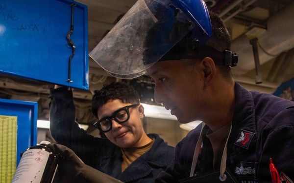 Two U.S. Navy personnel are working in what appears to be a shipboard laboratory or chemical handling space. The foreground individual is wearing a face shield, black rubber gloves, and a Navy-marked coverall while handling a dark chemical bottle. A second person in the background observes while holding open a blue storage cabinet.