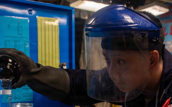 A technician wearing a blue face shield and black rubber gloves is carefully pouring liquid from a dark bottle into a graduated cylinder in what appears to be a laboratory setting. A bottle labeled 'NITRO' with a 'DATE OF PREP' label is visible in the foreground. The setting appears to be an industrial or military laboratory environment based on the blue equipment panel and signage visible in the background.