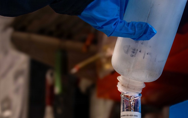 A person wearing blue nitrile gloves is pouring liquid from an inverted plastic bottle into a graduated glass cylinder. The setup appears to be a controlled liquid measurement procedure in what looks like a laboratory or technical workspace. A beaker and printed documentation are also visible on the work surface.