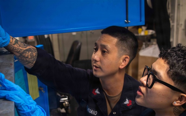 Two U.S. Navy personnel are working together in what appears to be a shipboard workspace or maintenance area. One sailor, identified by a name tape reading 'RIANO' and wearing U.S. Navy insignia, appears to be supervising or instructing the other. Both are wearing blue nitrile gloves and dark Navy coveralls while handling equipment near a blue storage cabinet.