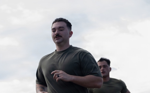 A group of military personnel in matching olive green PT (physical training) uniforms are running together on what appears to be a tarmac or flight deck. The lead runner is a young male with a mustache wearing a black ring and Adidas athletic shoes. The formation suggests an organized unit physical fitness run.