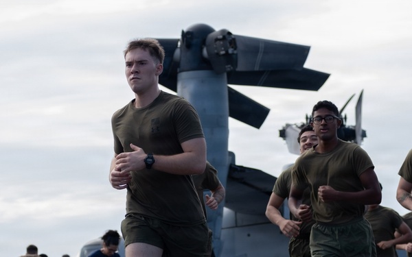 A group of military personnel in olive drab PT (physical training) uniforms are running on what appears to be the flight deck of a naval vessel. Behind them, a large tiltrotor aircraft — identifiable as a V-22 Osprey — is parked on the deck with its rotors folded. Additional personnel in the background appear to be conducting other activities on the flight deck.