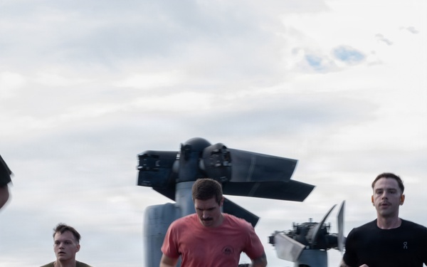 Military personnel are conducting a physical fitness run on the flight deck of a naval vessel. In the background, a V-22 Osprey tiltrotor aircraft is parked on the deck with its rotors folded. Multiple service members in PT (physical training) attire are participating, with observers standing nearby.