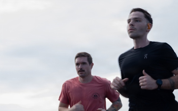 A group of people are running outdoors on what appears to be a large flat surface, possibly a ship's flight deck or a wide open platform, with an overcast sky in the background. Two men are prominently featured in the foreground — one wearing a red Quiksilver t-shirt and black shorts, the other in a black 'On' brand athletic shirt with visible leg tattoos. Several other runners are visible in the background, suggesting an organized group run or fitness event.