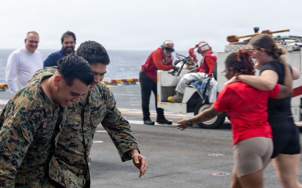 The image shows what appears to be a military homecoming reunion on the flight deck of a naval vessel. Marines in camouflage uniforms are running toward civilians, with joyful expressions suggesting an emotional greeting after a deployment. The scene takes place on an aircraft carrier or amphibious assault ship deck, with deck equipment and crew members visible in the background.