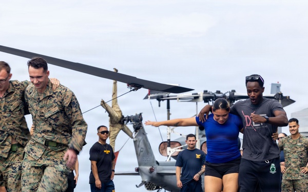 Military personnel and civilians participate in what appears to be a three-legged race or partner relay activity on the flight deck of a naval vessel. Two pairs of participants are actively racing while a crowd of onlookers cheers from the sides. A helicopter is visible in the background, confirming the flight deck setting.