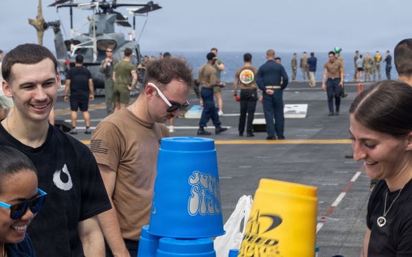 Military personnel are gathered on the flight deck of a naval vessel participating in a recreational activity involving large 'Jumbo Stacks' cup-stacking sets in blue and yellow. The event appears to be an organized morale or recreation day, with numerous service members visible in the background engaged in various activities. A sailor with the name tag 'Cortez' wearing a blue uniform is prominently featured alongside others in casual military attire.