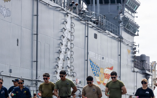 A group of military personnel and civilians are gathered on a pier or flight deck alongside a large gray naval vessel, participating in what appears to be a recreational or morale event. A man in a blue flight suit is speaking into a microphone, seemingly hosting or announcing an activity. Participants appear to be holding small objects, possibly eggs or balls, suggesting a team game or competition.