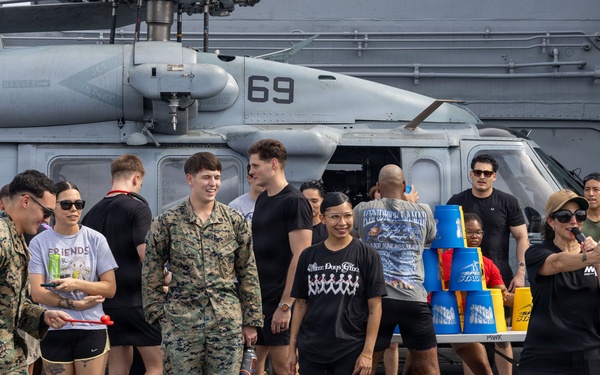 Military personnel and civilians are participating in a recreational event on the flight deck of a naval vessel, with a helicopter marked '69' visible in the background. The gathering appears to be a morale, welfare, and recreation (MWR) activity featuring games and social interaction. A mix of uniformed Marines in camouflage and civilians in casual attire are engaged in what appears to be a cup-stacking or carnival-style game.