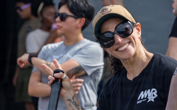 A smiling woman wearing a black 'MWR' t-shirt, tan baseball cap, and large round sunglasses poses for the camera while holding a microphone. She has visible tattoos on her forearm and is making a peace/victory sign gesture. Several other people are visible in the background in an outdoor setting.