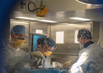 A surgical team of at least three medical professionals is performing an operation in what appears to be a hospital operating room. The image is captured through a gap in blue surgical draping, giving a framed view of the procedure in progress. The team is wearing full sterile surgical attire including gowns, gloves, masks, and surgical caps.