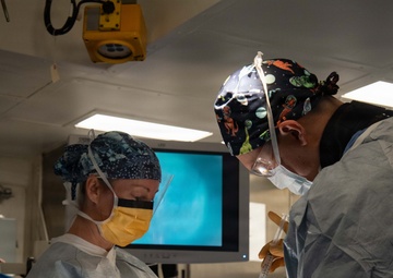 Three medical professionals are performing a surgical procedure in an operating room. The team appears to be actively engaged in a procedure, with one surgeon using a syringe while another holds surgical instruments. A monitor displaying what appears to be an endoscopic or imaging feed is visible in the background.