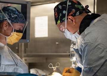 Two medical professionals are performing a surgical procedure in an operating room. The surgeon on the right is actively working with a hemostat/clamp instrument on the surgical site, while the assistant on the left observes and assists. Both are wearing standard surgical personal protective equipment including gowns, gloves, masks, and surgical caps.