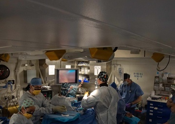 A surgical team of approximately 7-8 medical personnel is performing an operation in what appears to be a shipboard operating room, indicated by the low ceiling, structural markings, and compact layout. The team is gathered around a patient on an operating table covered with blue surgical draping, with a laparoscopic monitor visible in the background displaying a live surgical feed. The environment suggests a military or naval medical facility based on the room's construction and labeling visible on the wall.